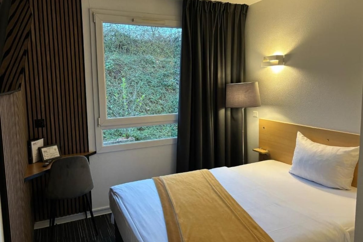 Ein Hotelzimmer mit Bett, Schreibtisch, Stuhl, Fenster und einer Wandlampe.