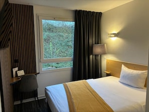 Ein Hotelzimmer mit Bett, Schreibtisch, Stuhl, Fenster und einer Wandlampe.