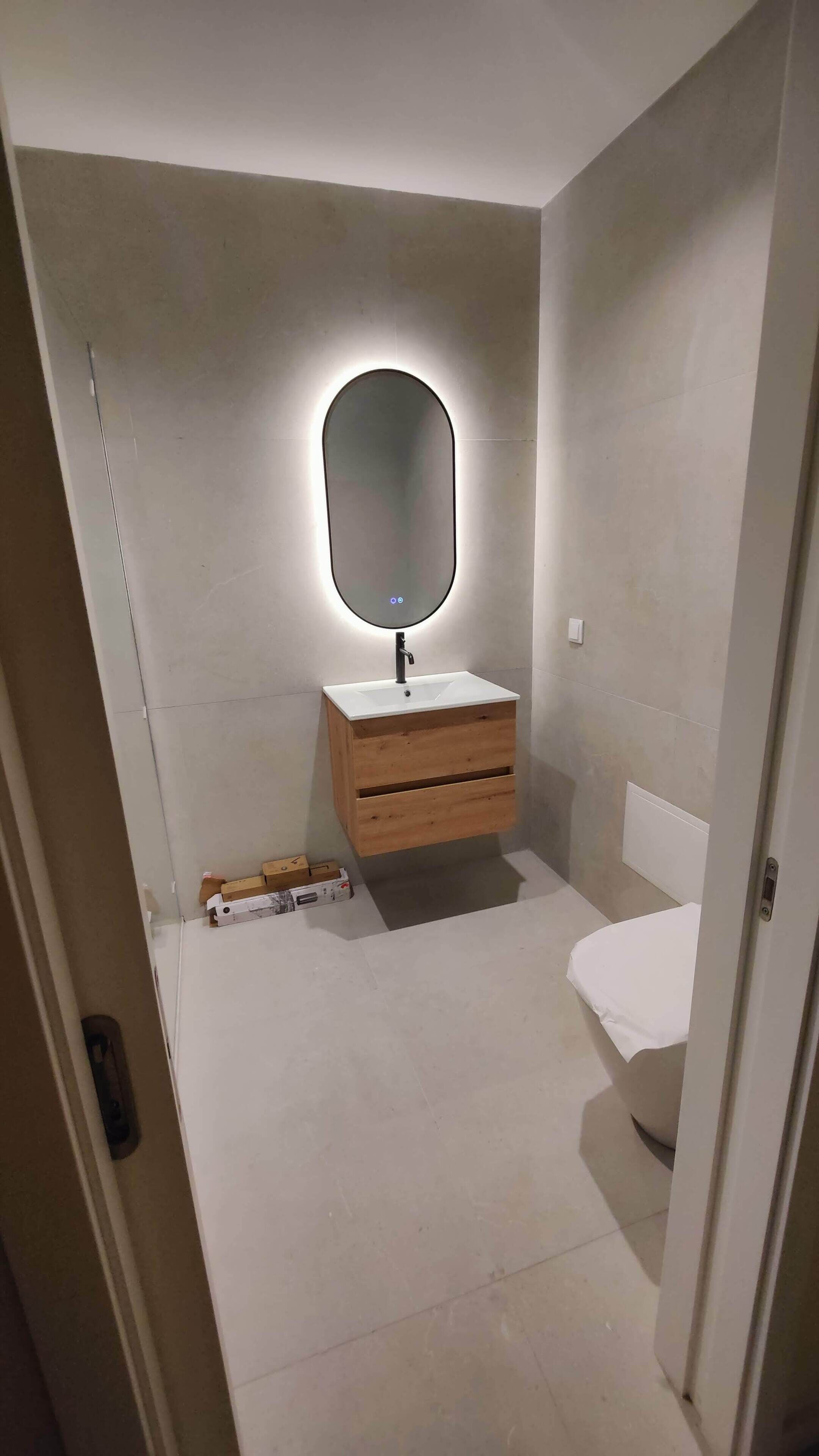 Een kleine badkamer met een witte wastafel, een houten wastafelmeubel en een spiegel met een ronde verlichting.