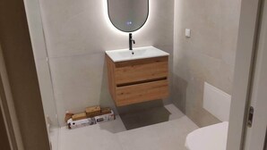 Uma casa de banho pequena com uma pia branca, toucador de madeira e um espelho com luz circular.