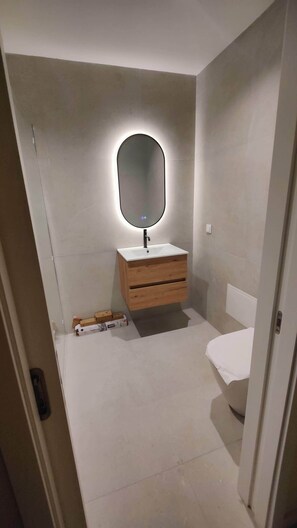 Ein kleines Badezimmer mit einem weißen Waschbecken, einer Holzkommode und einem Spiegel mit runder Beleuchtung.