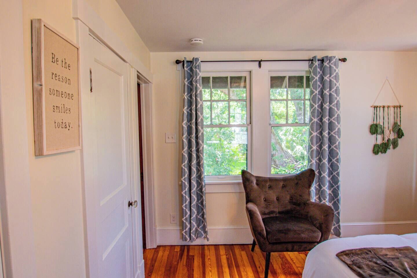 A room with a wooden floor, a chair, a bed, a window with patterned curtains, and a wall-mounted macrame decoration.