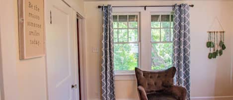 A room with a wooden floor, a chair, a bed, a window with patterned curtains, and a wall-mounted macrame decoration.