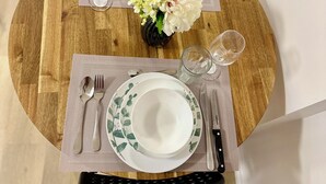 Un tavolo da pranzo rotondo in legno con una tovaglia bianca e verde a motivi, un vaso con fiori bianchi e un set di posate e bicchieri.