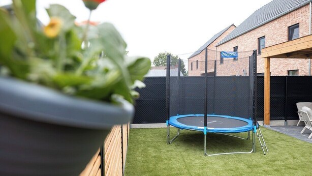 Een achtertuin met kunstgras, een trampoline, een houten omheining en een gebouw met een bord.