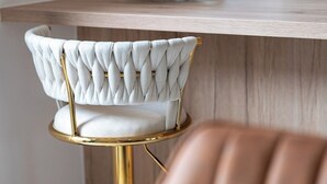 Een barstoel met een beklede zitting en gouden frame, een witte fauteuil met een geborduurde rugleuning, en een houten bar met een glazen blad.