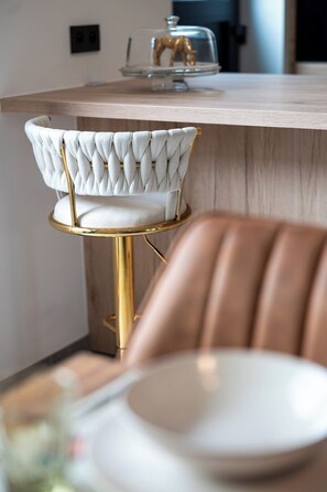 Ein Barhocker mit gepolstertem Sitz und goldfarbener Rahmen, ein weißer, gepolsterter Stuhl mit gebuckelter Rückenlehne sowie eine Holzelemente-Bar mit einer Glasplatte.