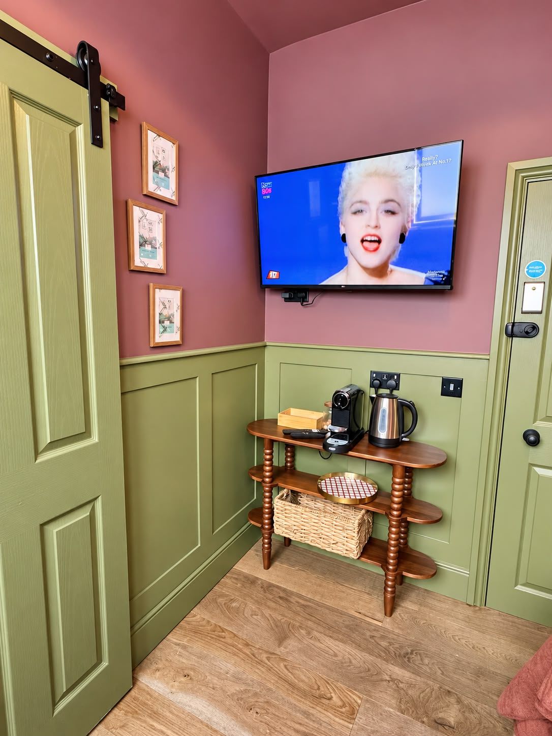 A room with a television mounted on the wall displaying a news channel, a wooden table with a kettle and a camera, and a door with a black handle.