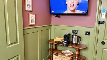 A room with a television mounted on the wall displaying a news channel, a wooden table with a kettle and a camera, and a door with a black handle.