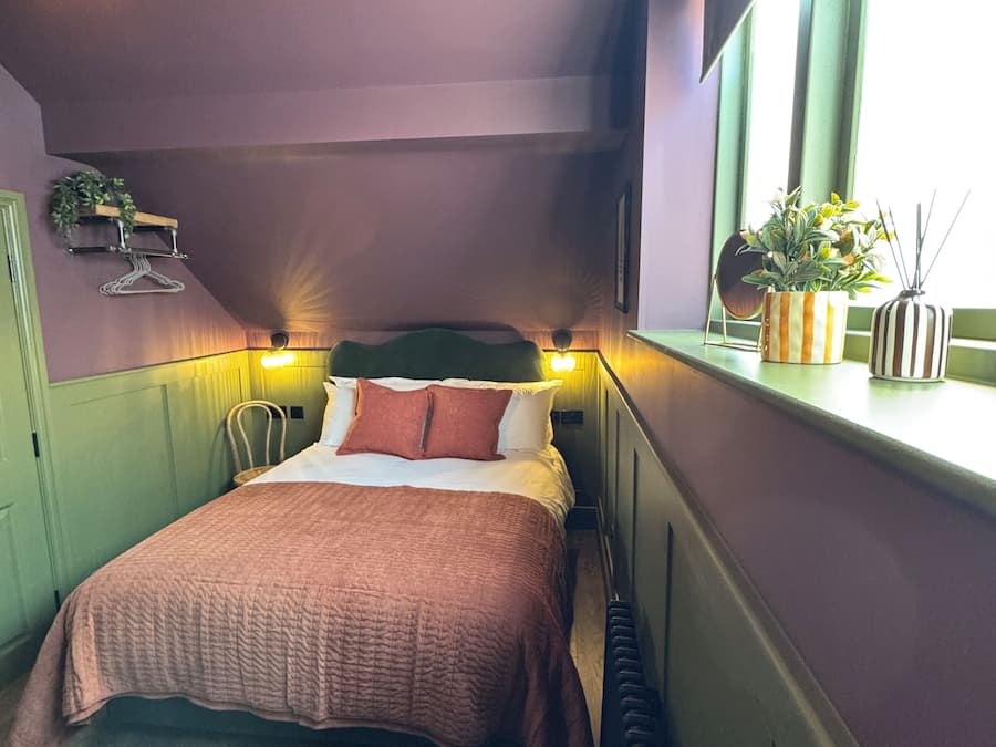 A bedroom with a bed, a window with plants, a hanging light fixture, and a radiator.