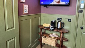 A room with a television mounted on the wall, a wooden table with a kettle and a box, and a door with a handle. - The Linen Rooms (Knutsford)