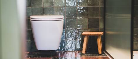 A toilet with a white lid, a small wooden stool, and a tiled wall with a reflective surface.