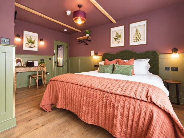 A bedroom with a large bed, a desk, and a chair. There are framed pictures on the wall, a hanging plant, and a ceiling light.