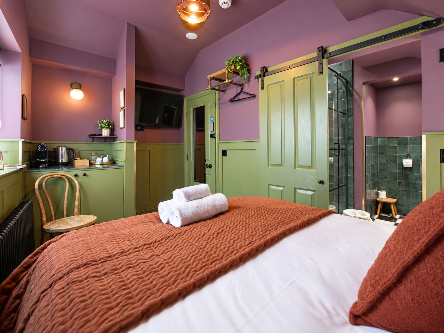 A hotel room with a bed, a chair, a small table, a green door, a shower area, and a kitchenette.