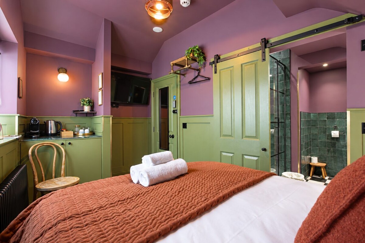 Habitación de hotel con cama, una silla, una mesita, una puerta verde, zona de ducha y kitchenette.