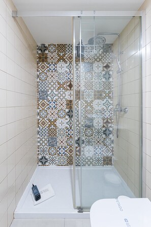 A shower with a glass door, a white bathtub, and a tiled wall with a geometric pattern. - GuestReady-Cosy Studio with Balcony in Porto (Porto)