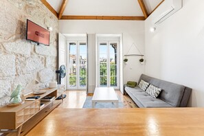 A modern living room with a stone wall, wooden beams, a grey sofa, and a flat-screen TV. - GuestReady-Cosy Studio with Balcony in Porto (Porto)