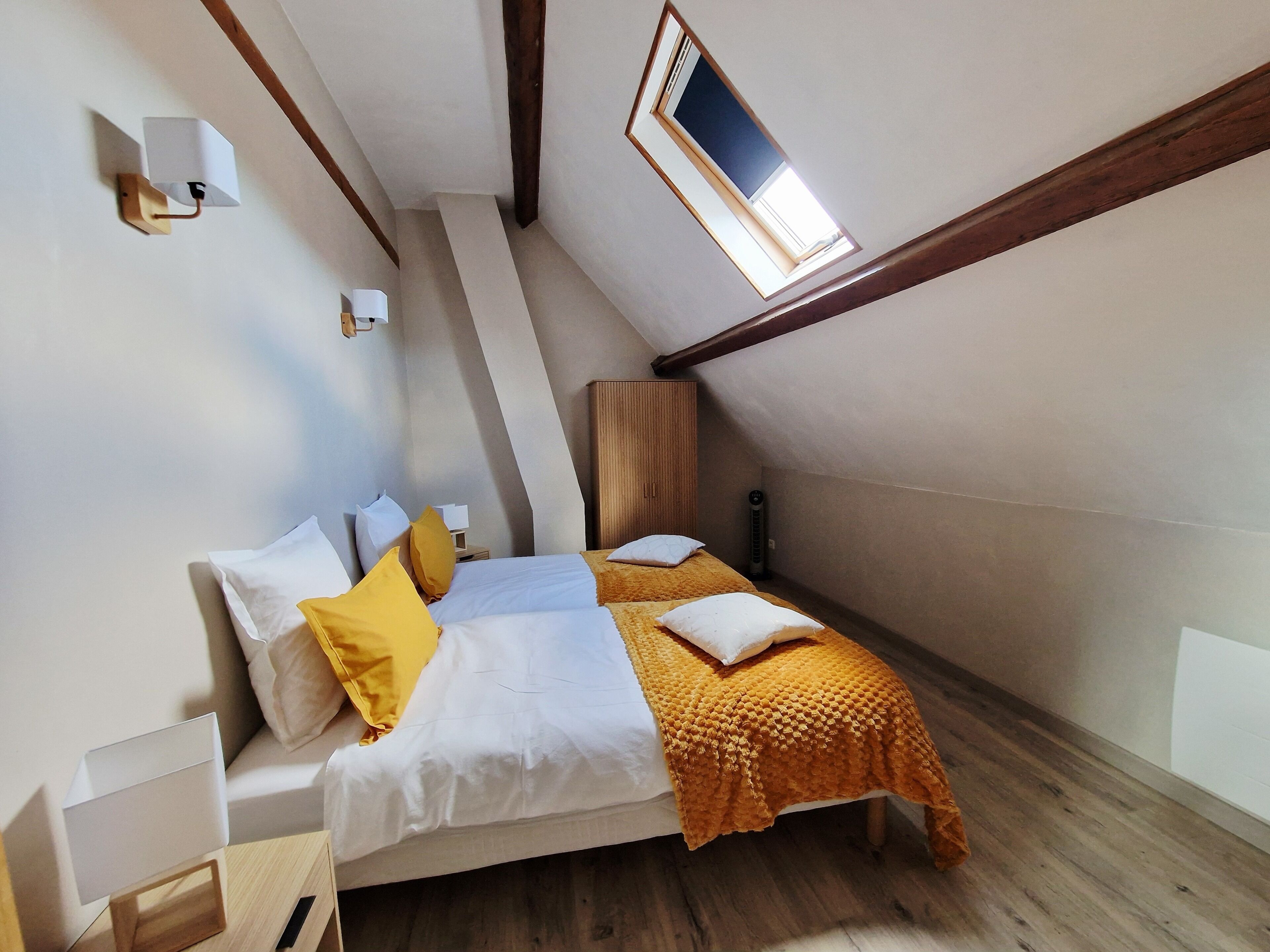 Une chambre avec un plafond en pente, un puits de lumière, un lit avec une literie blanche et des oreillers jaunes, une armoire en bois et une table de chevet.