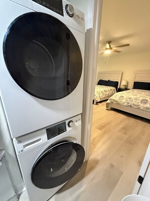 A compact, modern laundry room with a front-loading washer and dryer, a ceiling fan, and a bedroom with two beds. - The Seashell Sanctuary, Poolside, Sleeps 8, Beach, Boat Slip, cable & wifi (Madeira Beach)