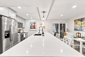 A modern kitchen with a large island, stainless steel appliances, and a dining area with white chairs and a table. - The Seashell Sanctuary, Poolside, Sleeps 8, Beach, Boat Slip, cable & wifi (Madeira Beach)