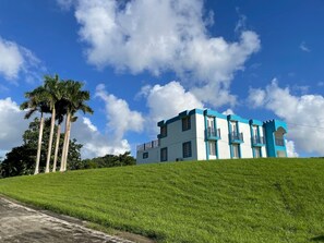 Ein zweistöckiges Gebäude mit blauer und weißer Fassade, das auf einem grasbewachsenen Hügel mit Palmen im Hintergrund steht.