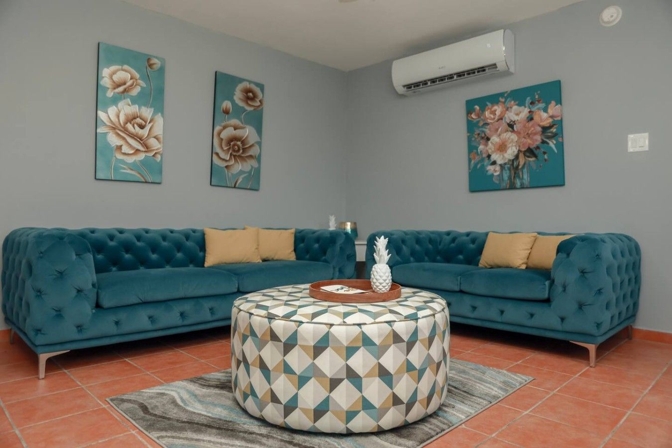 Un salon avec un canapé bleu, un pouf à motifs et des tableaux floraux accrochés au mur.