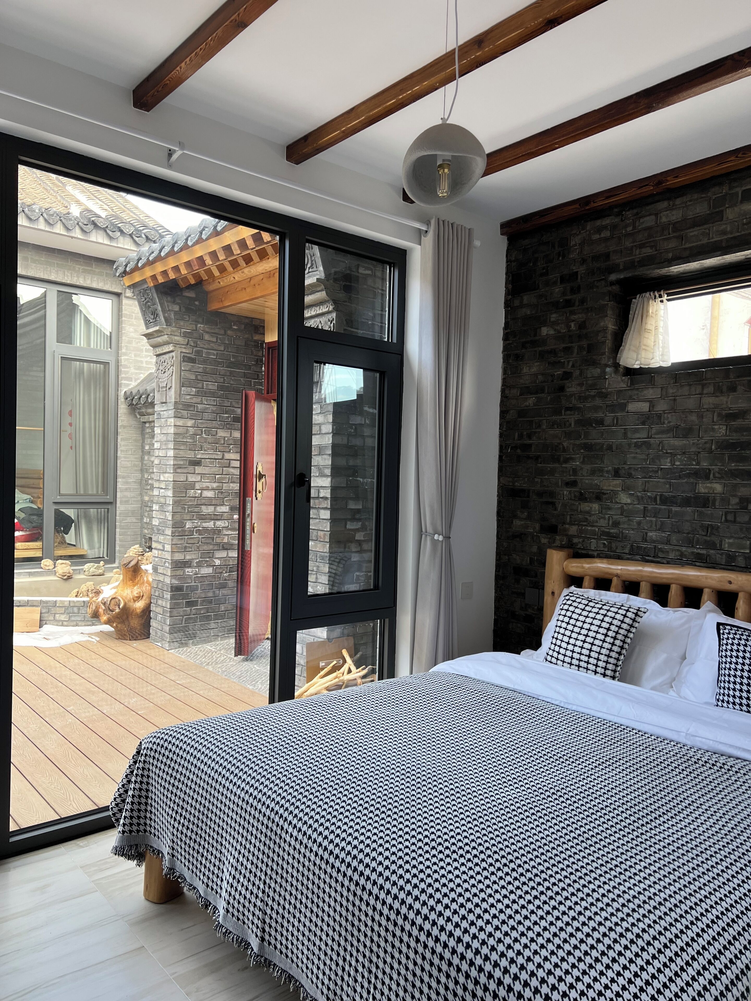 Een slaapkamer met een groot bed, een houten hoofdeinde, een zwart bakstenen muur en een schuifdeur van glas die leidt naar een buitenruimte.