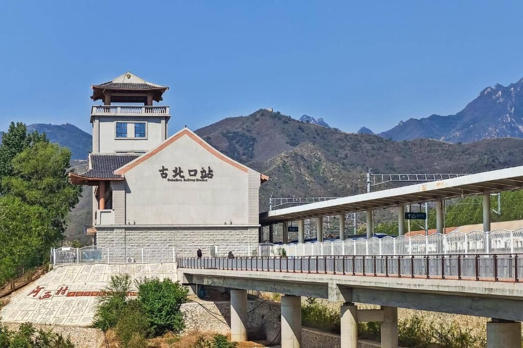 A station building with a sign, a pedestrian bridge, and mountains in the background.