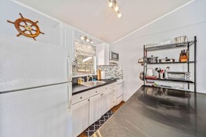 Una cocina con armarios blancos, encimera negra, decoración de timón de barco, salpicadero de azulejos y estante metálico con utensilios de cocina.