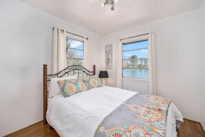 Um quarto com uma cama, duas janelas, uma mesinha de cabeceira e um abajur.