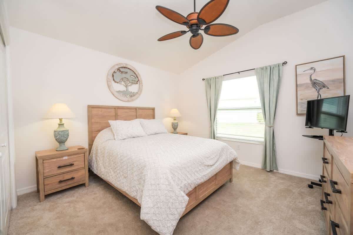 A bedroom with a bed, nightstands, a ceiling fan, a window with curtains, a television, and a wall-mounted artwork.