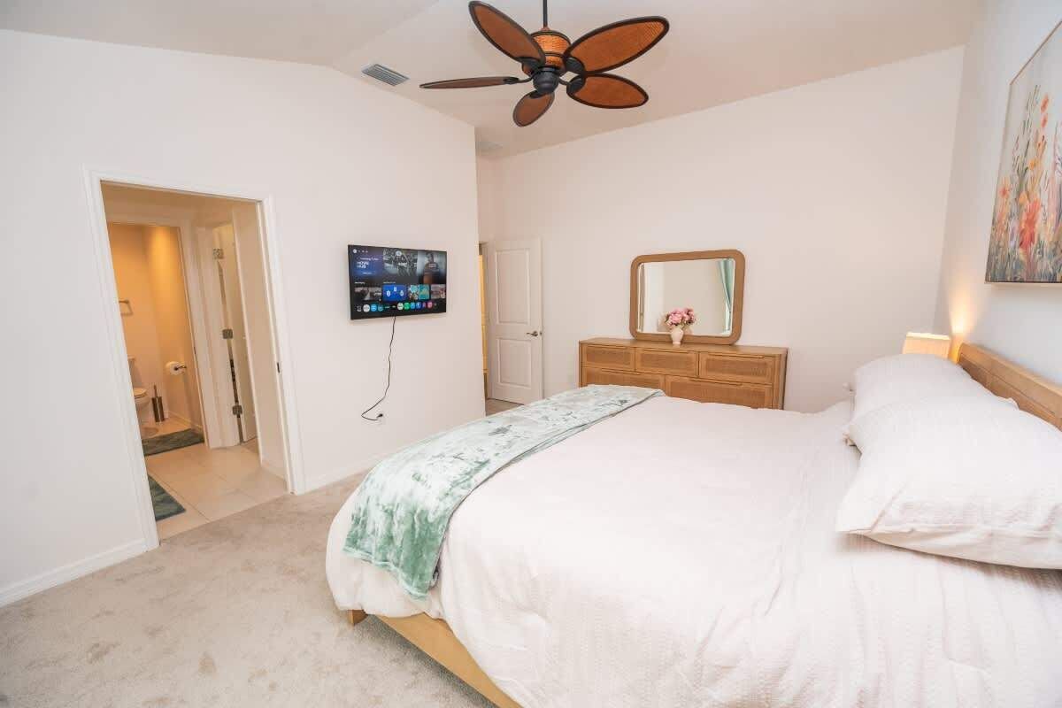 A bedroom with a bed, a ceiling fan, a television mounted on the wall, and a wooden dresser.