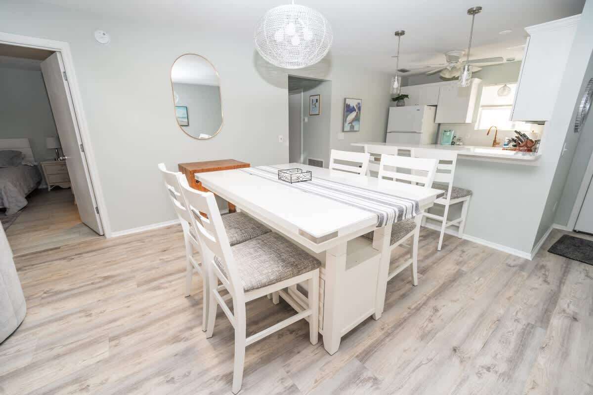 A dining area with a white table and chairs, a hanging light fixture, and a kitchen in the background.