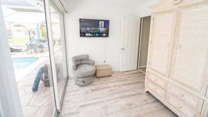 Ett rum med utsikt över poolen, en grå fåtölj, en tv som är monterad på väggen och en stor vit garderob.