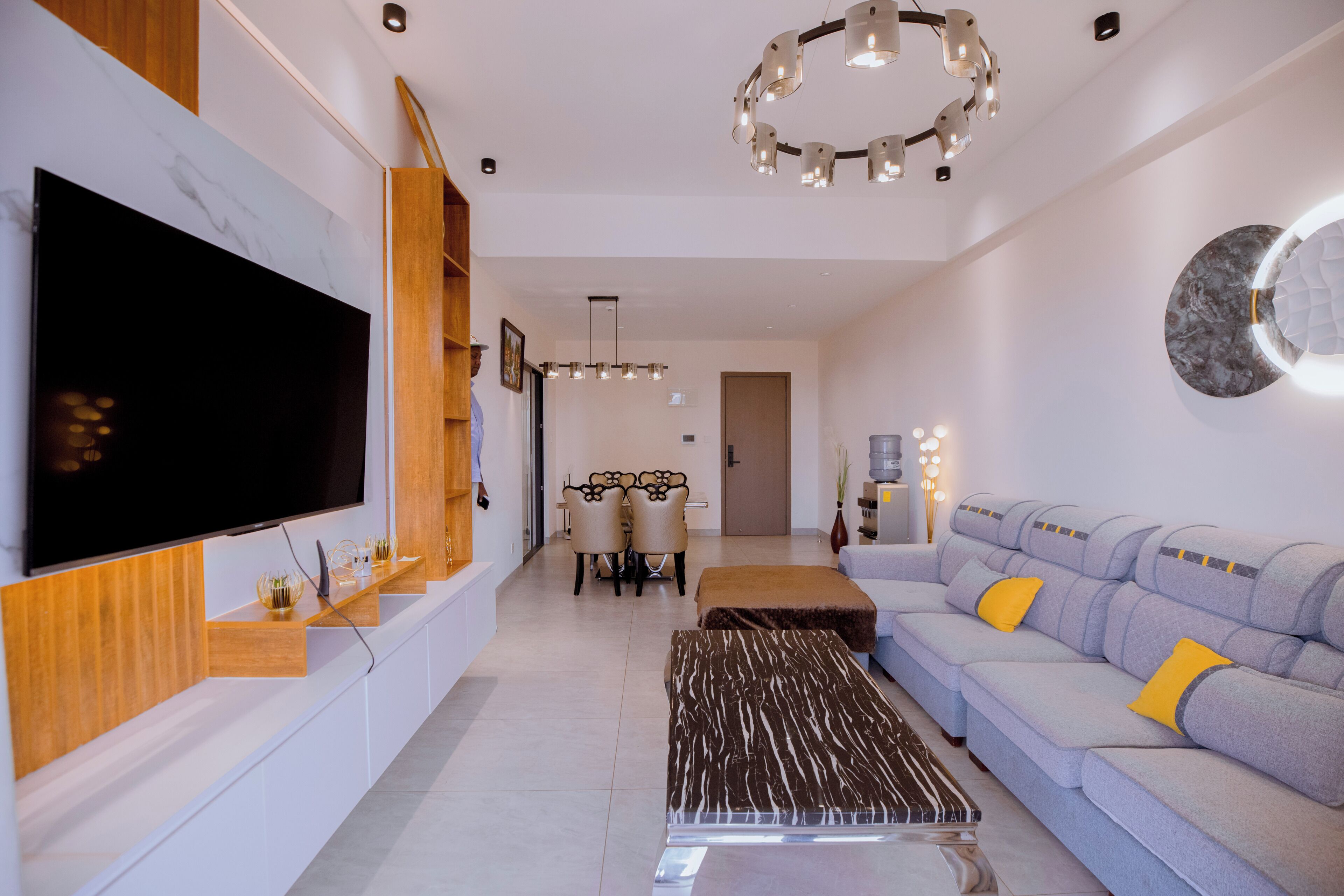 Een moderne woonkamer met een flatscreen televisie, een bank met kussens, een salontafel en een eethoek met stoelen.