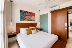 A hotel room with a bed, bedside tables, a ceiling fan, an air conditioning unit, and a colorful abstract painting on the wall. - Vibrant 2BR apartment (Playa del Carmen)