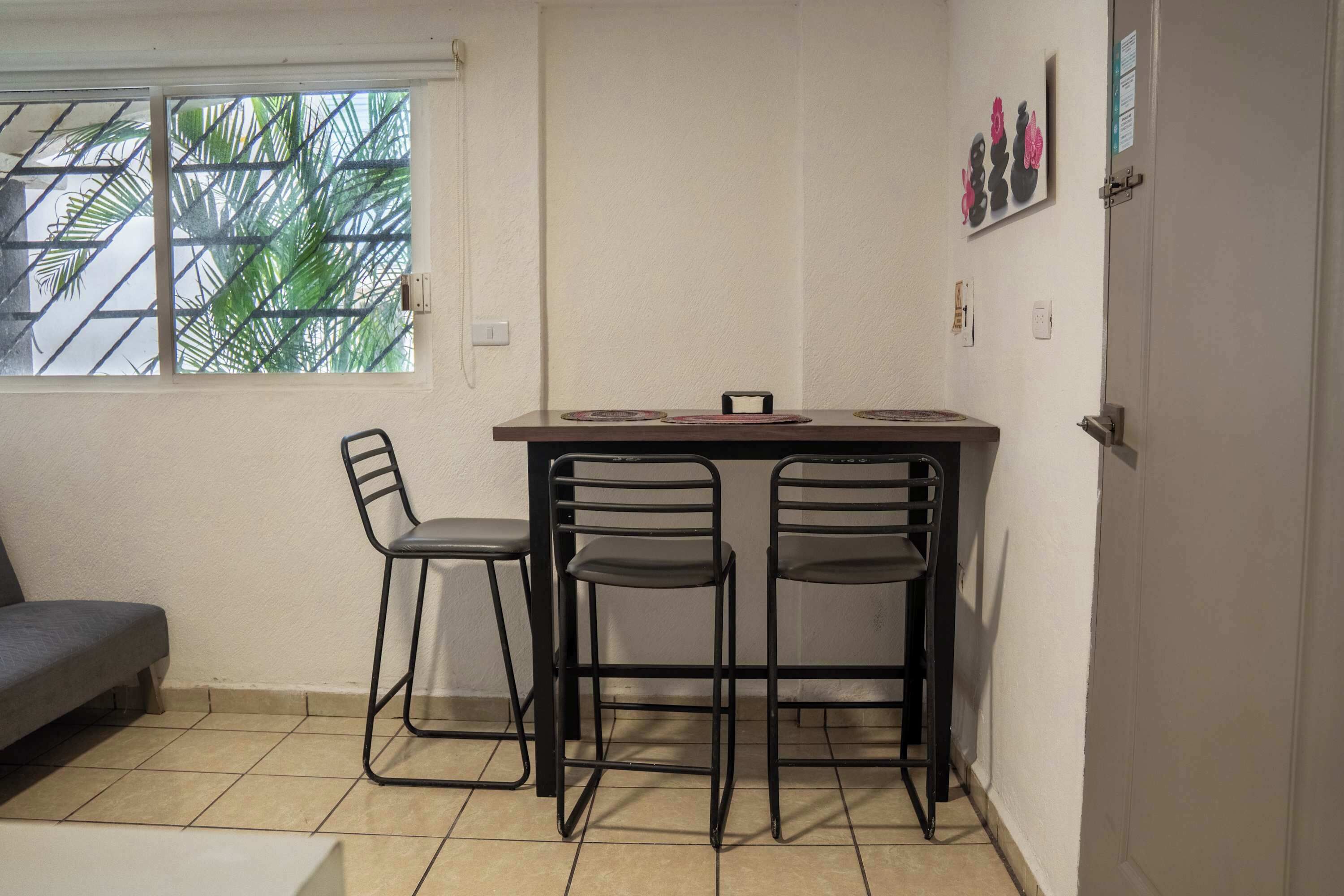 Una habitación pequeña y acogedora con una mesa de comedor y cuatro sillas. Hay una ventana que da a palmeras y una puerta a la derecha.