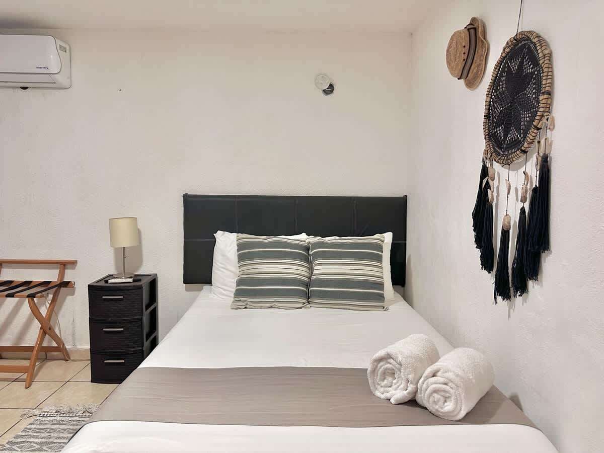 A neatly made bed with striped pillows, a bedside lamp, a small wooden chair, and a wall-mounted dreamcatcher.