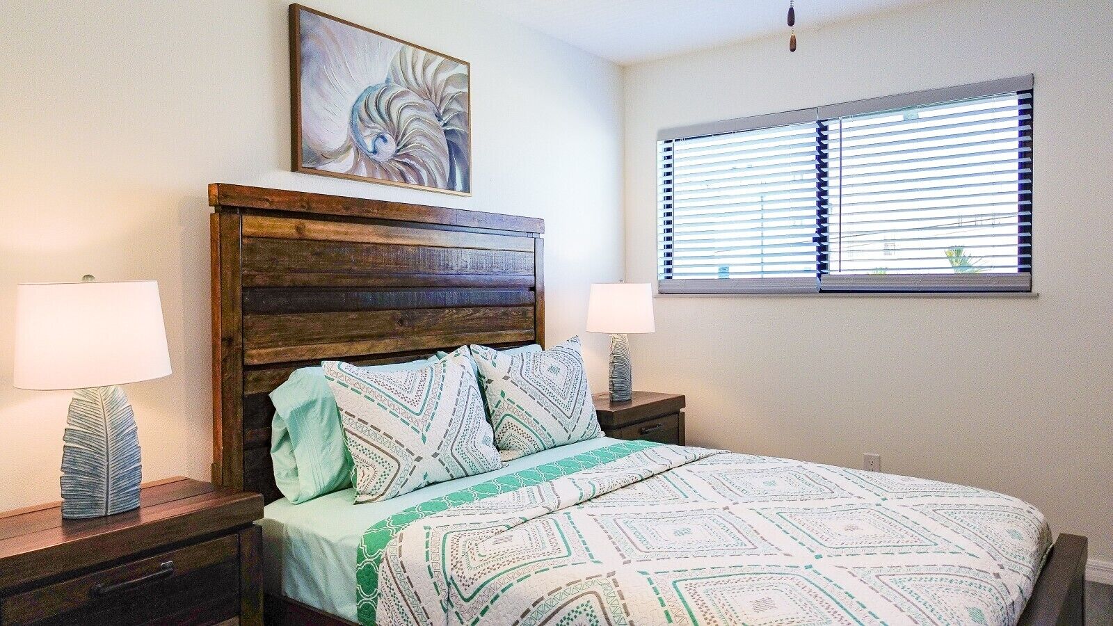 Une chambre à coucher avec une tête de lit en bois, une couvre-lit à motifs, deux lampes de chevet et une fenêtre avec des stores.