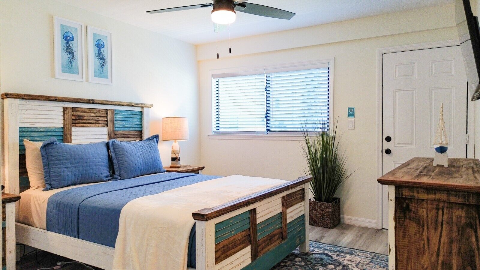A bedroom with a bed, a nightstand, a lamp, a window with blinds, a door, and a ceiling fan.