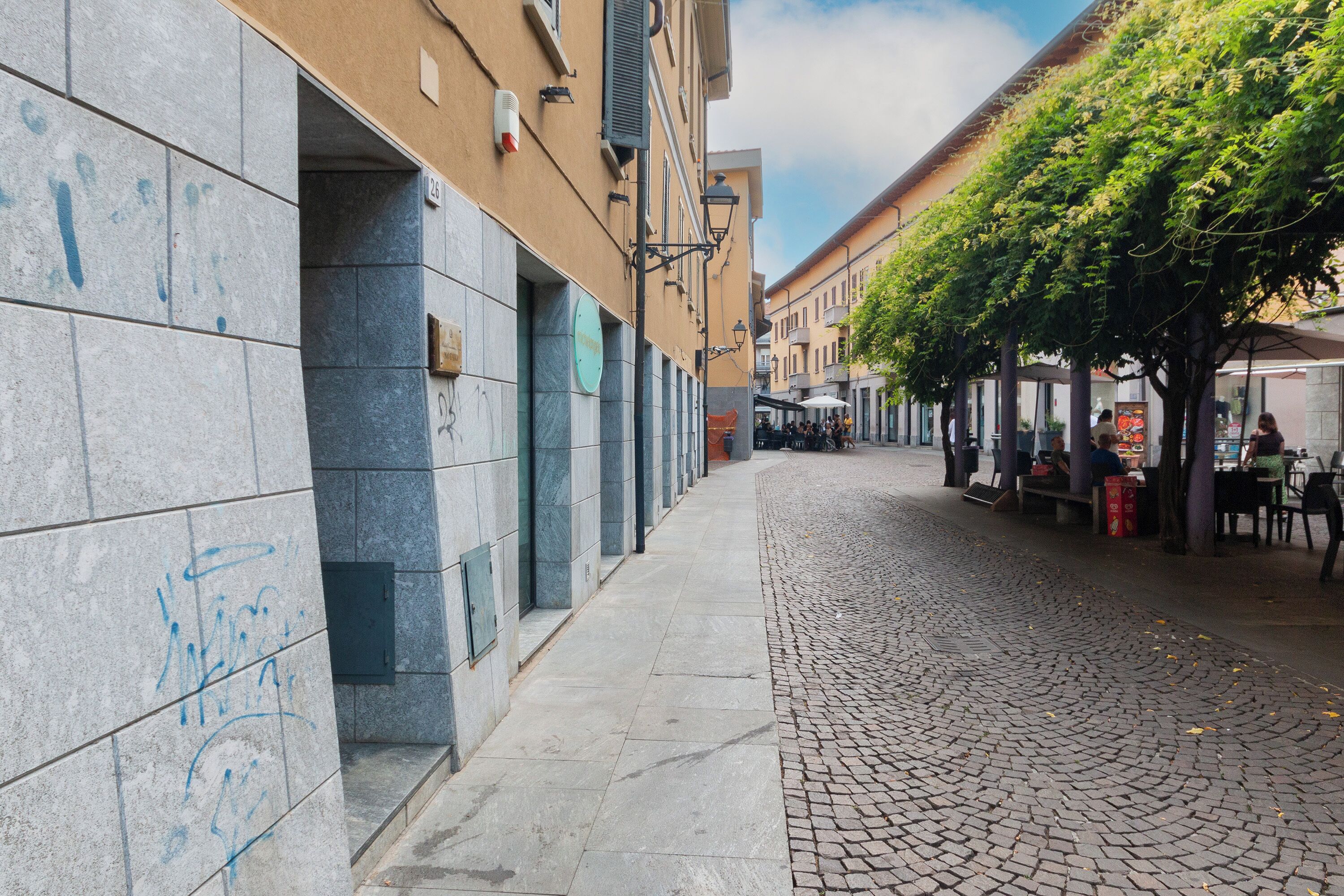 Una calle adoquinada con edificios a ambos lados, mesas al aire libre bajo árboles y un cielo despejado.