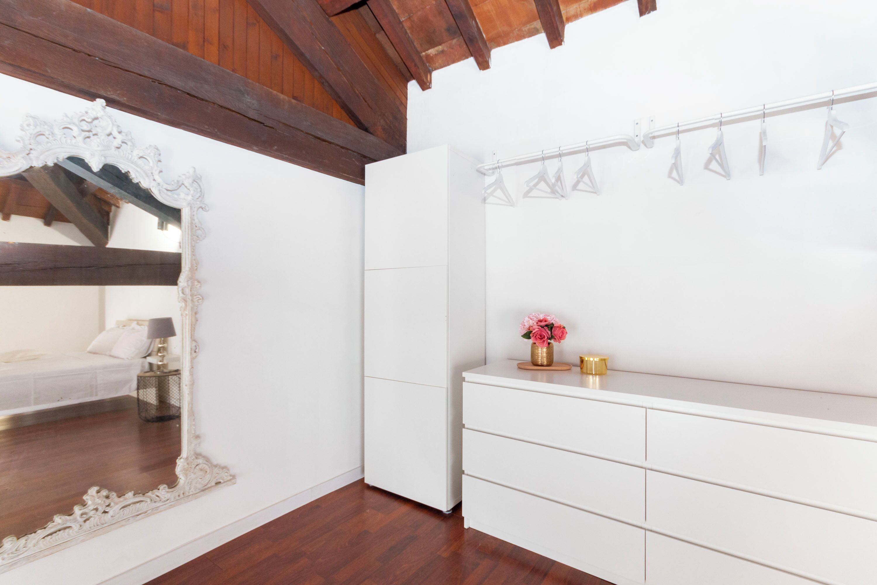 Ein Zimmer mit einem weißen Bett, einem großen verzierten Spiegel, einem weißen Kleiderschrank und einem kleinen Tisch mit Blumen in einer Vase.