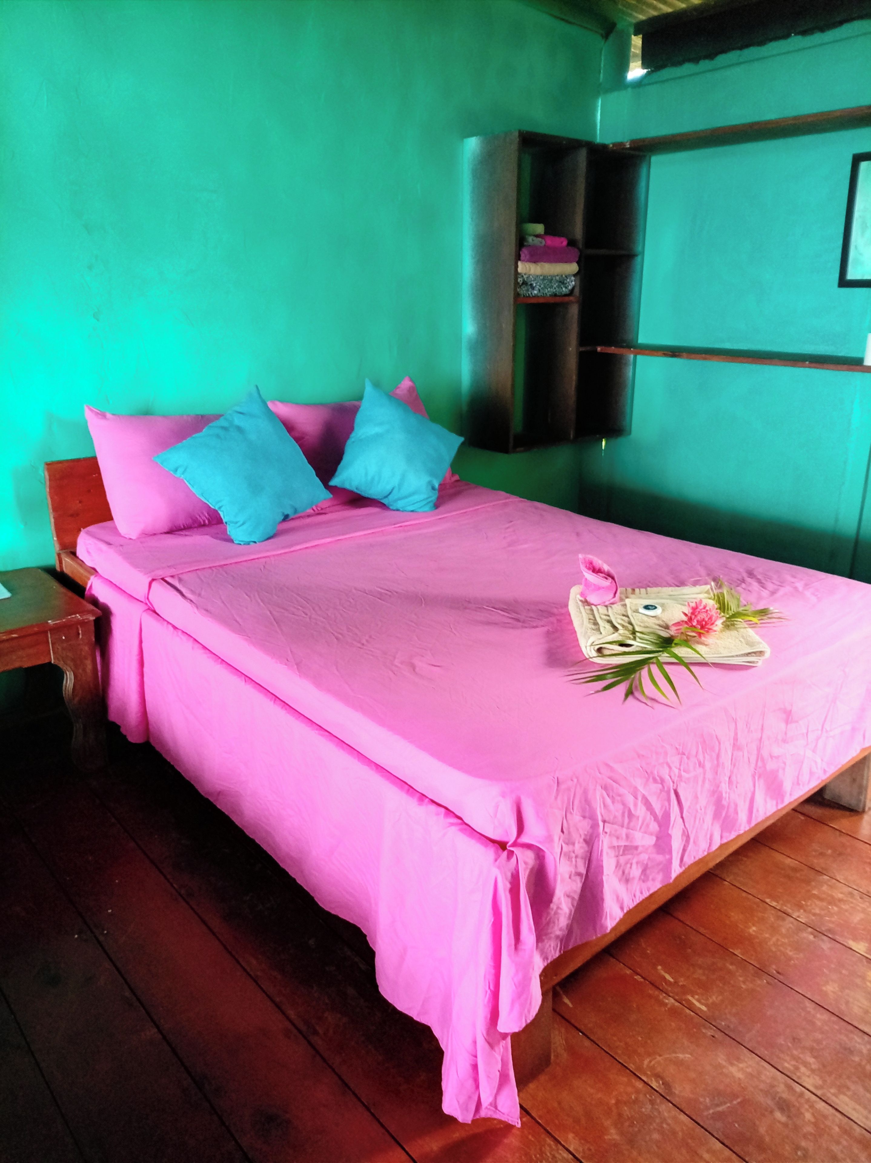 A bed with pink bedding and blue pillows, a wooden nightstand, and a small shelf with folded clothes.
