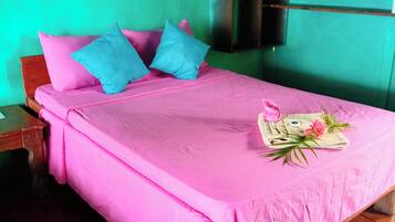Cama com roupa de cama rosa e almofadas azuis, mesa de cabeceira de madeira e uma pequena estante com roupas dobradas.