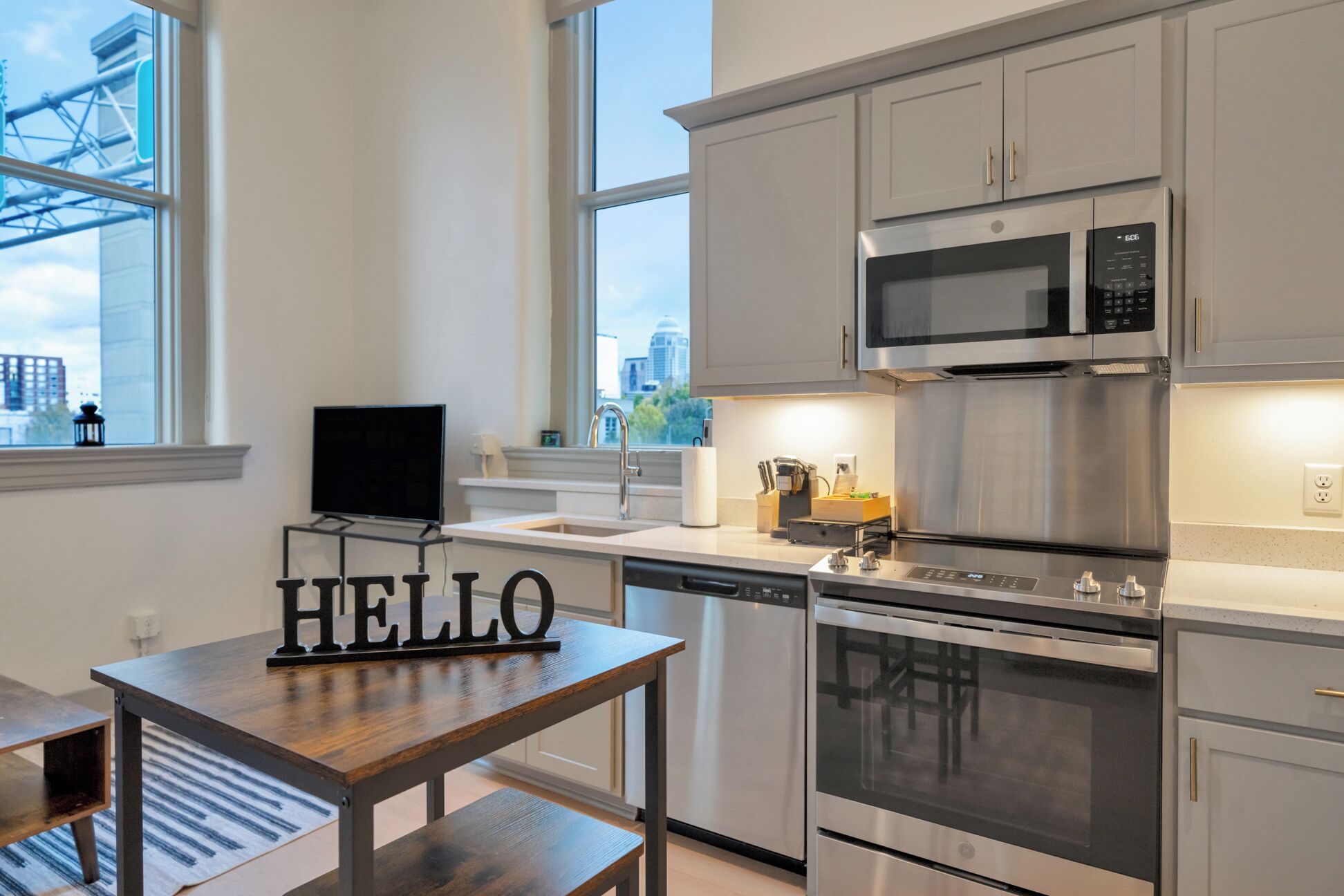 Une cuisine moderne avec des appareils en acier inoxydable, une table en bois ornée d’une inscription « HELLO », et une vue sur la ville par la fenêtre.