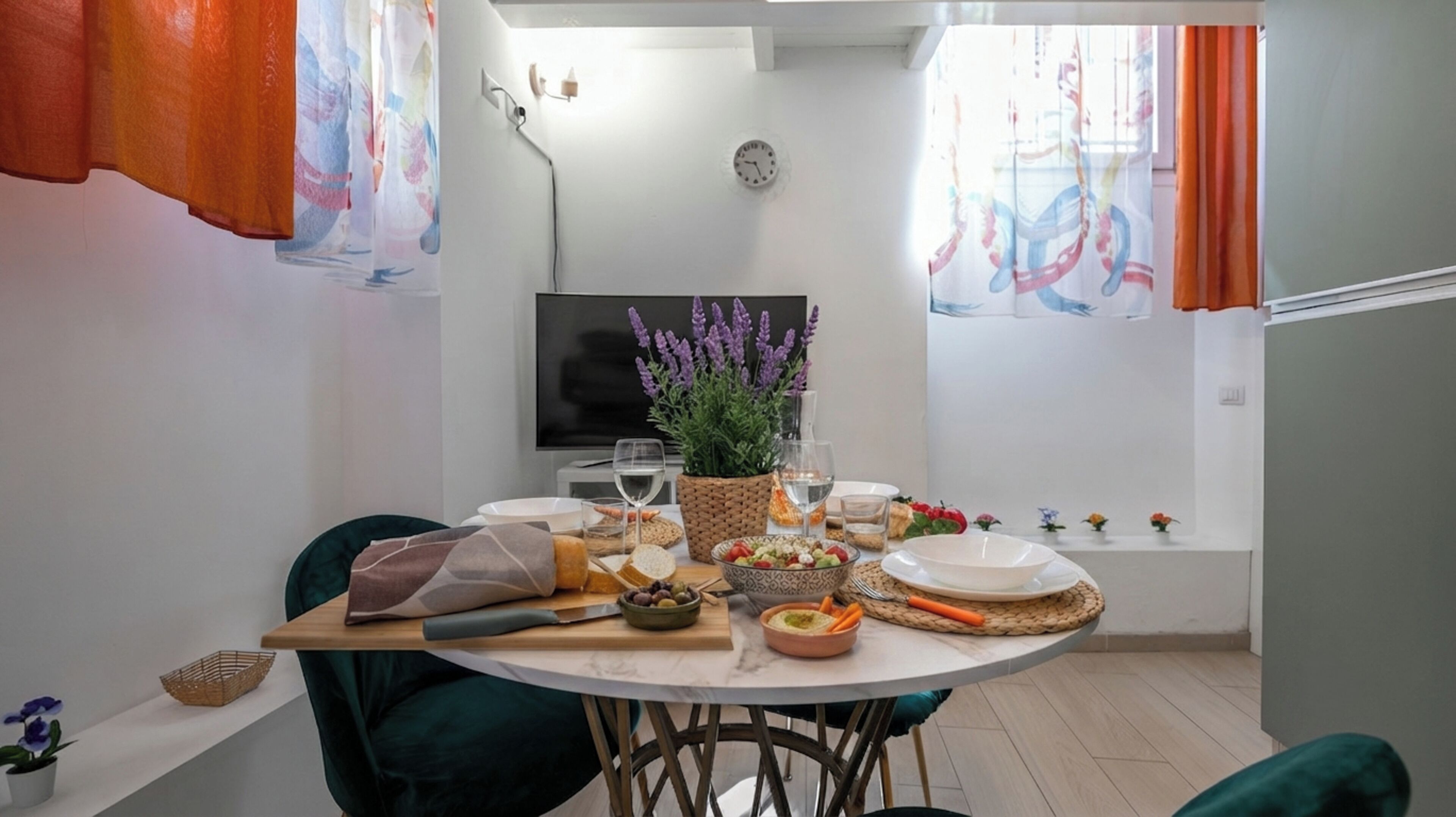 A dining area with a round table set for two, a television, and a window with curtains.