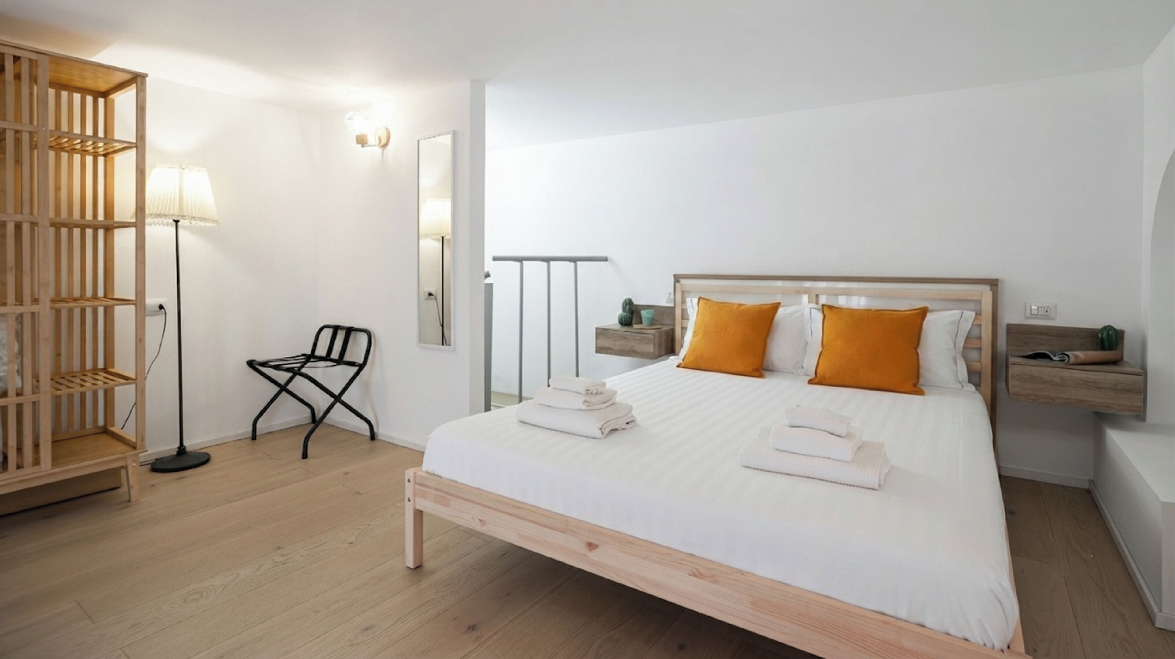 A bedroom with a bed, a chair, a floor lamp, a mirror, and a wooden shelf.