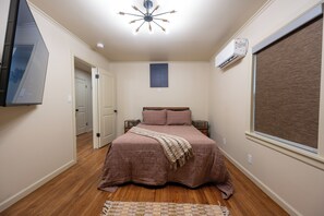 Ein Schlafzimmer mit einem Bett, einem Deckenventilator, einer Klimaanlage, einem Fenster mit Jalousien und einer Tür zu einem anderen Raum.