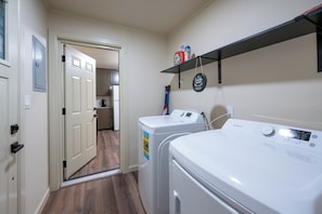 A laundry room with two front-loading washing machines, a shelf with laundry products, and a door leading to a kitchen area. - Luxury Apartment C on TU Campus and 2 miles to downtown (Tulsa)