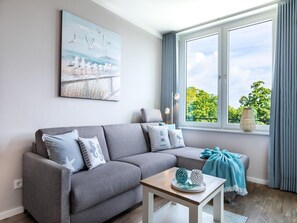 A modern living room with a grey sofa, a small table with decorative items, a painting of seagulls, and a large window with curtains. - Parkside Retreat in der Nahe von Baltic (Lübeck)